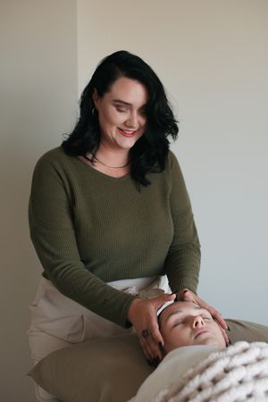 A person in a green sweater performs a facial massage treatment on a client lying on a white bed.