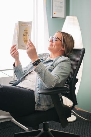 A person in glasses and denim jacket reclines in an office chair while reading a book and smiling.