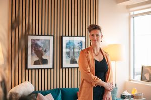Person in orange blazer and black top stands in modern living room with wood slat wall and framed artwork.