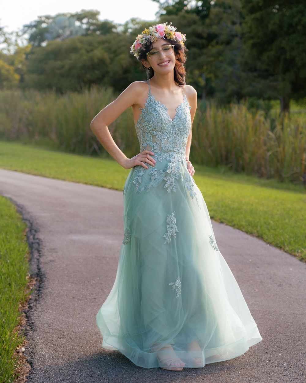 Young woman in sage green lace A-line gown with floral crown posing on outdoor pathway.