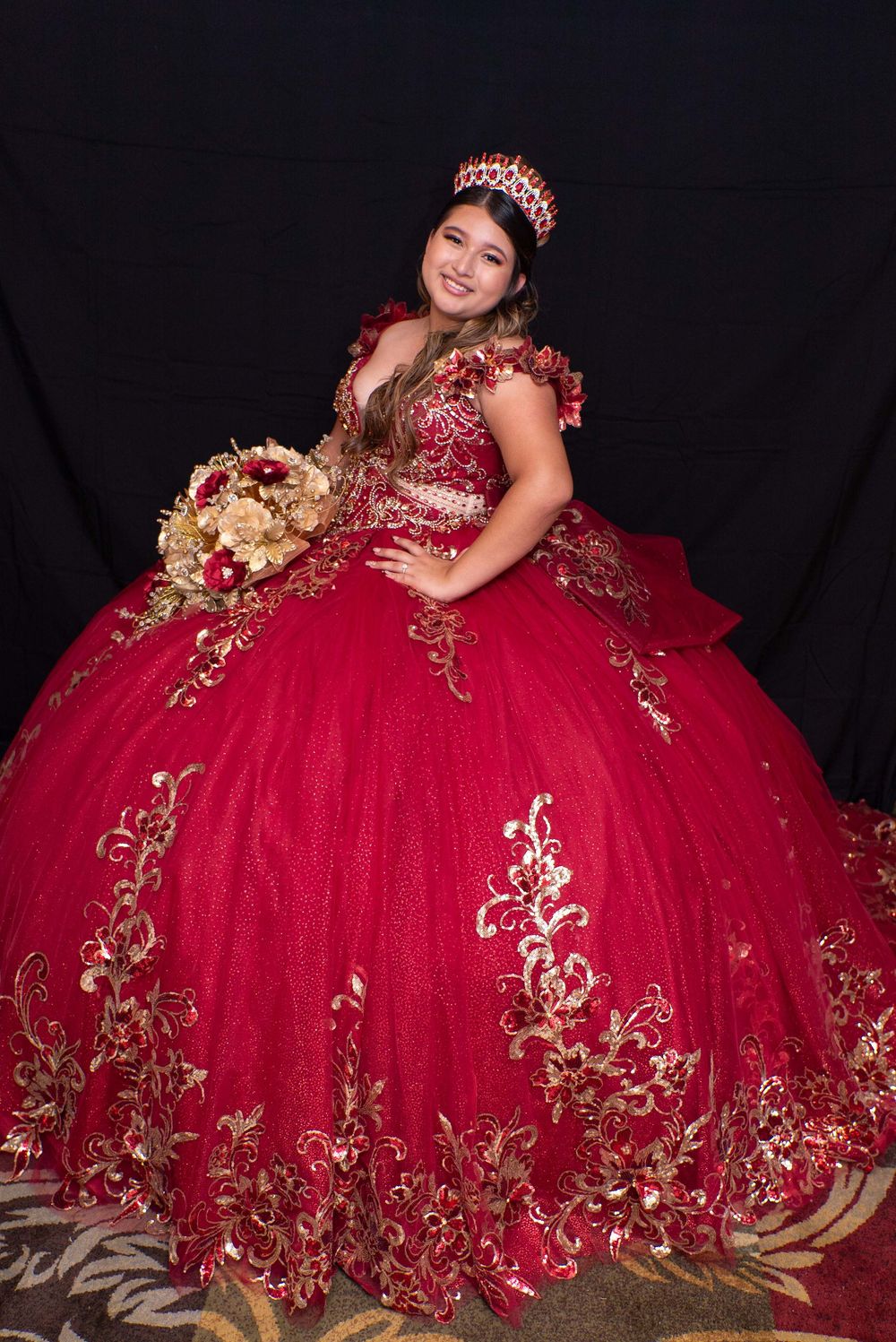 Young woman in red and gold quinceañera ball gown with crown and floral bouquet posing against black background.