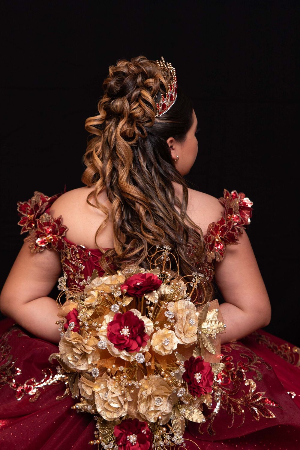 Back view of quinceañera hairstyle with braided updo, curls, red crown, and red gold bouquet in formal gown.
