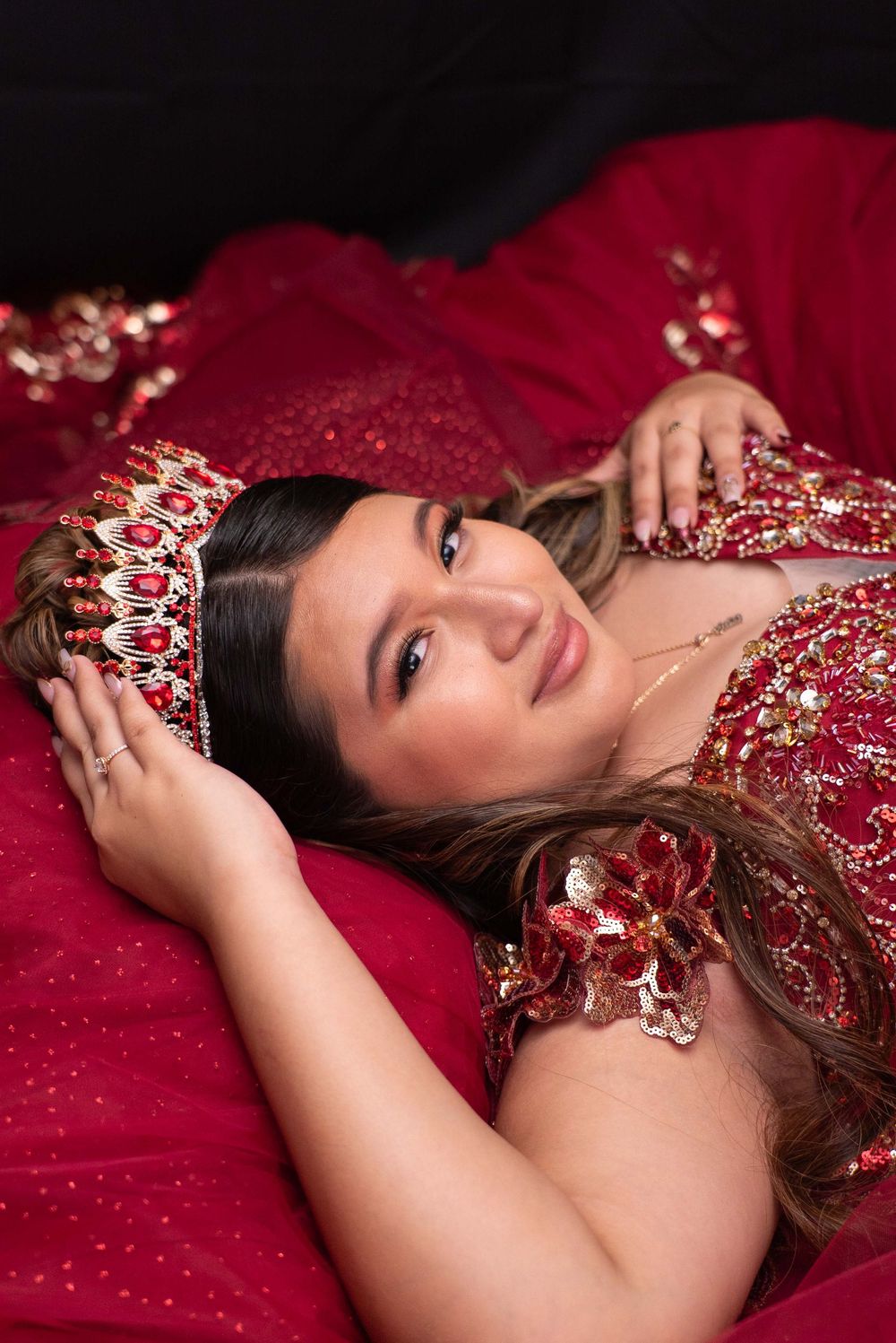 Young woman in red sequined quinceañera gown lying down holding jeweled crown, smiling at camera.