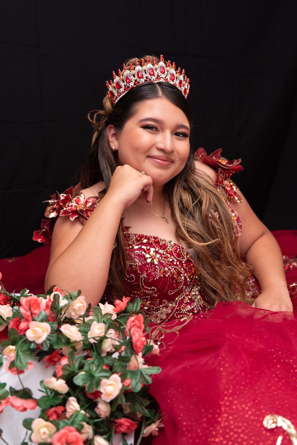 Smiling young woman in red beaded quinceañera dress and crown posing with pink roses in formal portrait.