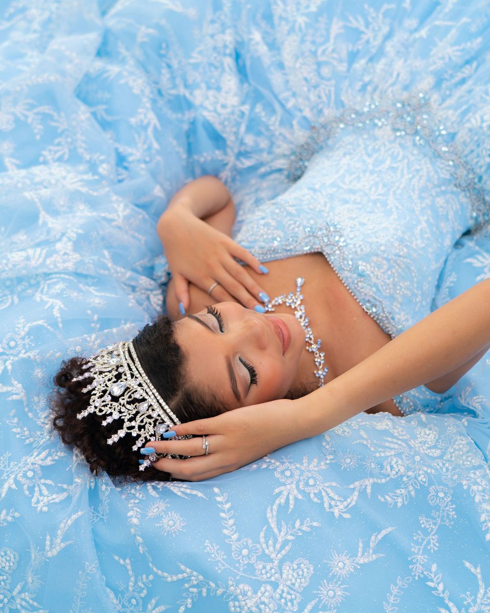 Young woman in light blue ball gown with crystal tiara and necklace lying gracefully among blue fabric.