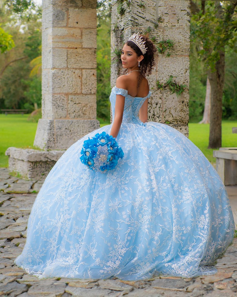 Elegant quinceañera in light blue lace ball gown holding blue bouquet standing between stone columns outdoors.