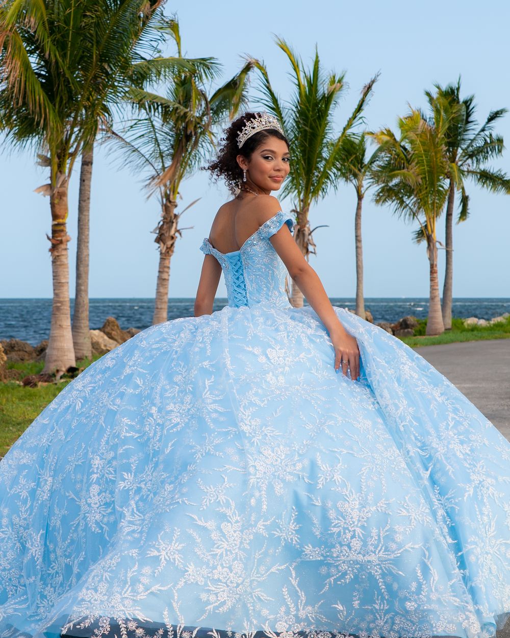 Young woman in light blue floral quinceañera ball gown with silver crown posing near palm trees by ocean.