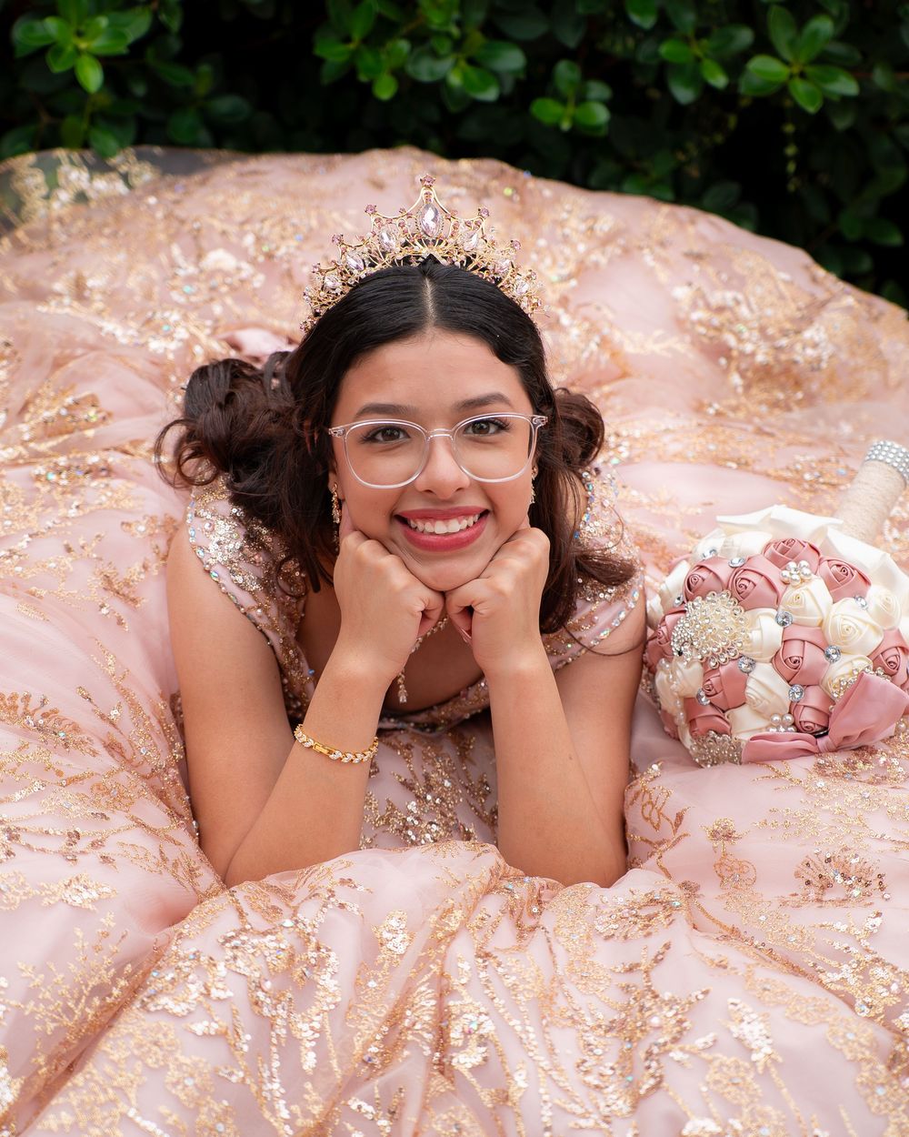 Smiling young woman in blush pink gold quinceañera gown with crown and glasses lying with floral bouquet.