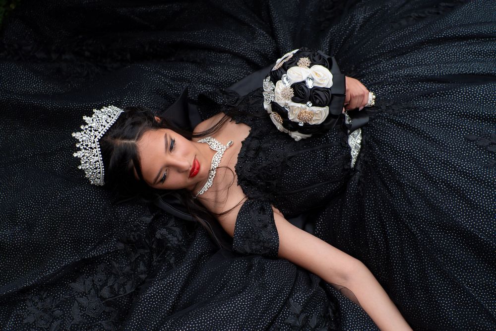 Young woman in black quinceañera ball gown with silver crown holding black and white bouquet, lying down.