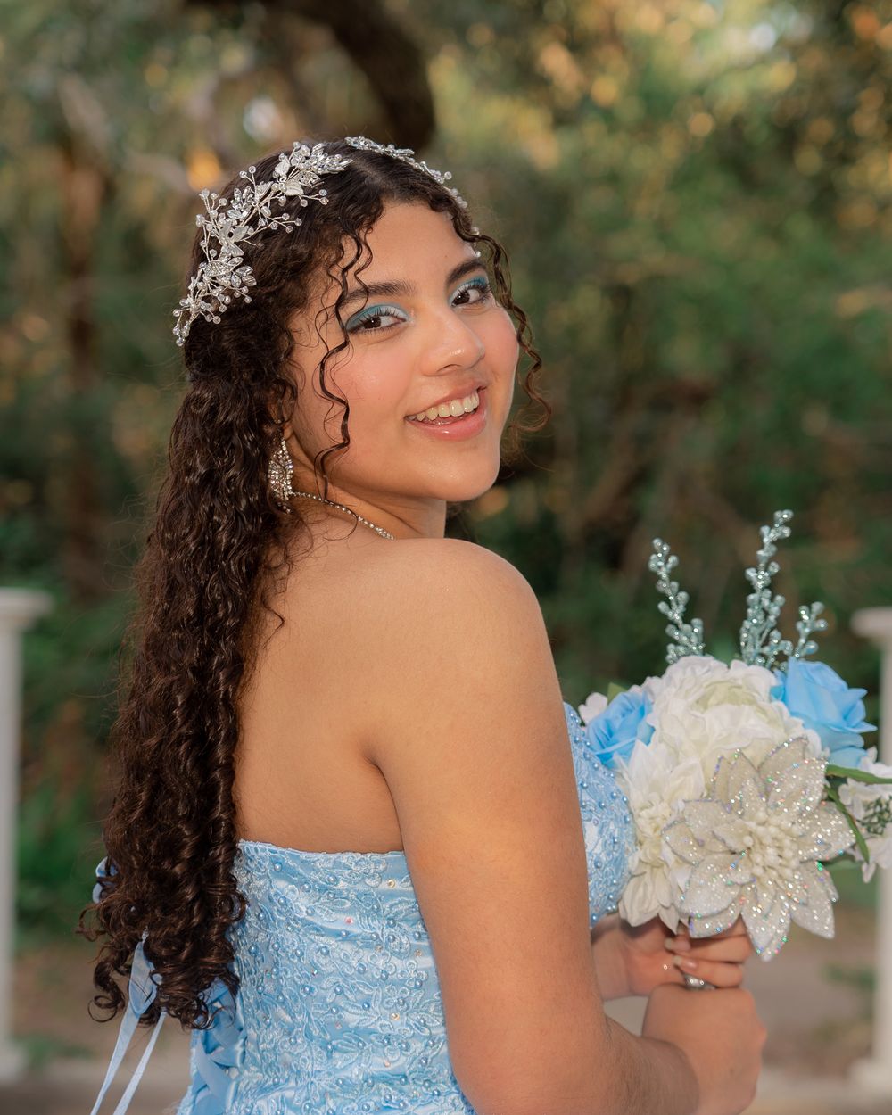Smiling young woman in blue quinceañera dress holding white bouquet wearing silver floral hair crown outdoors.