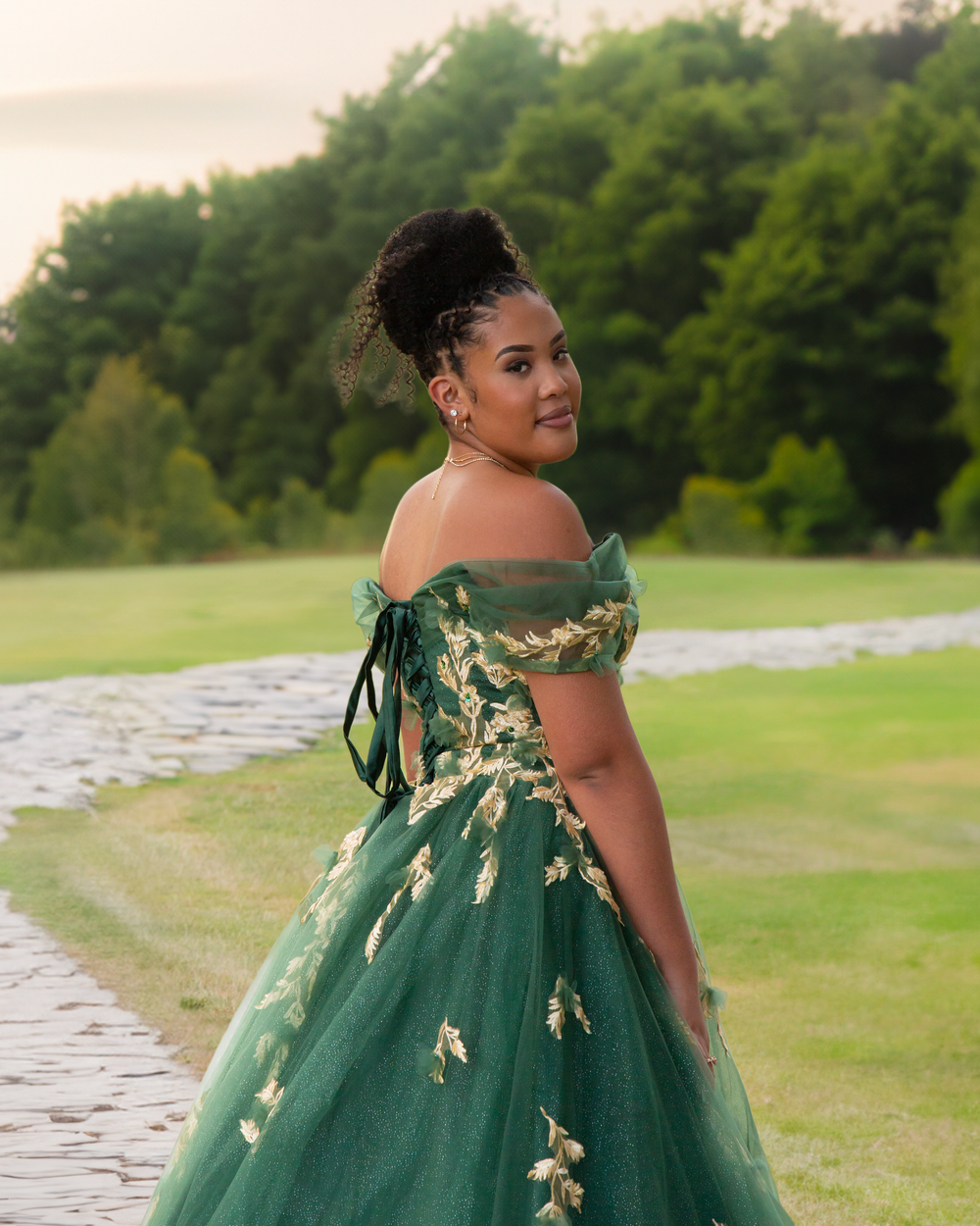 Young woman in emerald green off-shoulder quinceañera gown with gold details posing on outdoor stone path.