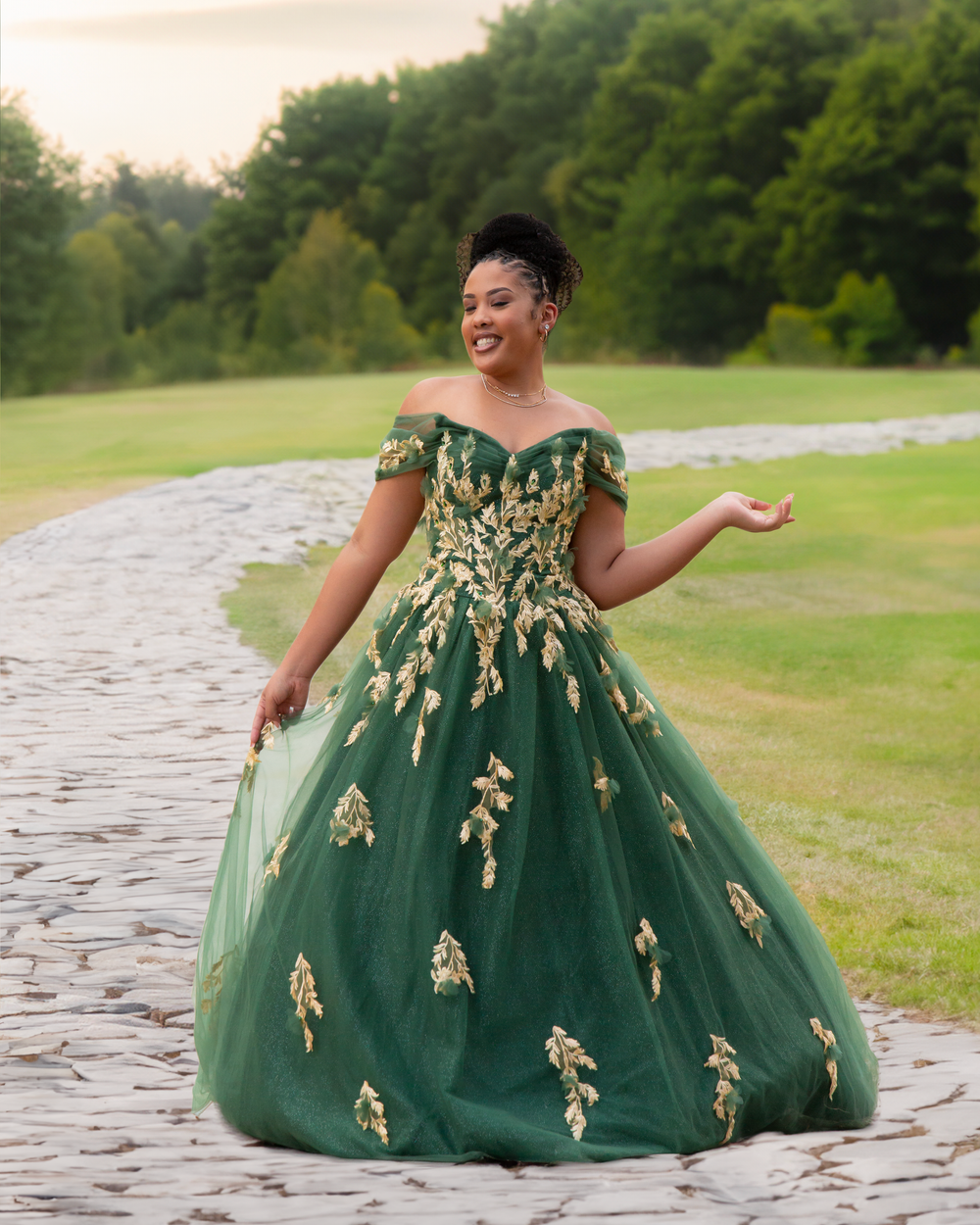 Young woman in emerald green off-shoulder ball gown with gold appliques posing on stone path outdoors.