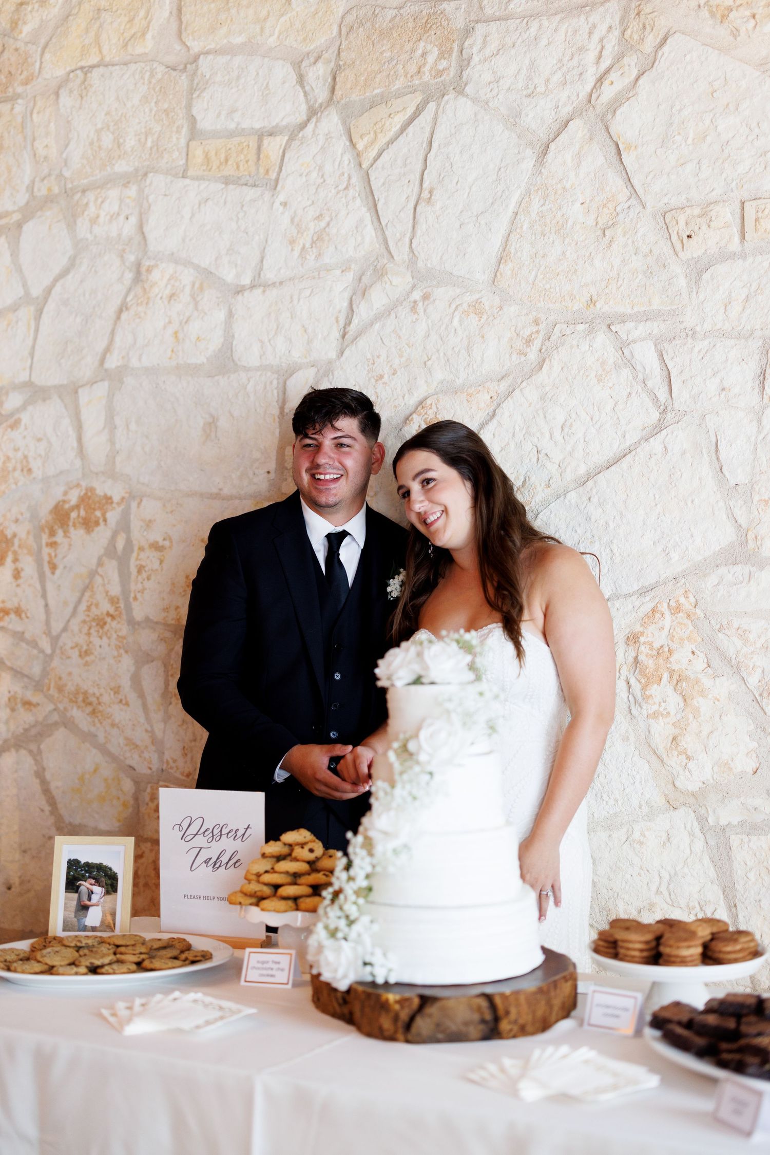 Wedding dessert table with white cake and cookies against a stone wall backdrop.
