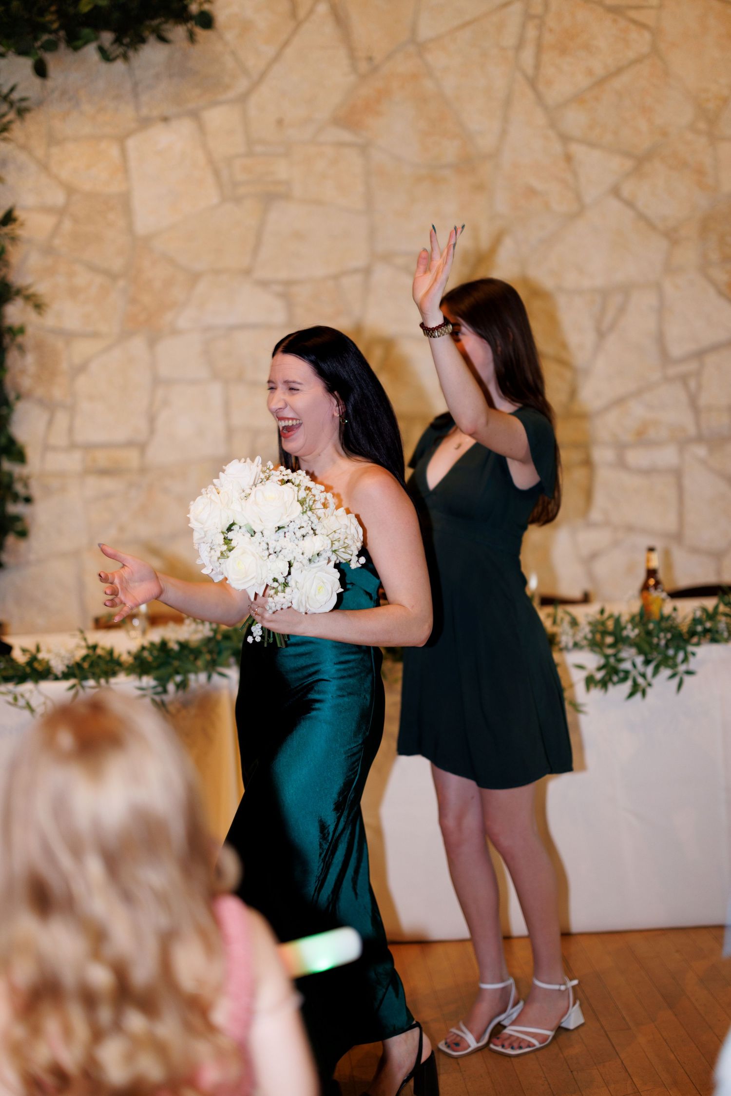 Two women dancing at a wedding reception, one holding a white bouquet in an elegant teal dress.