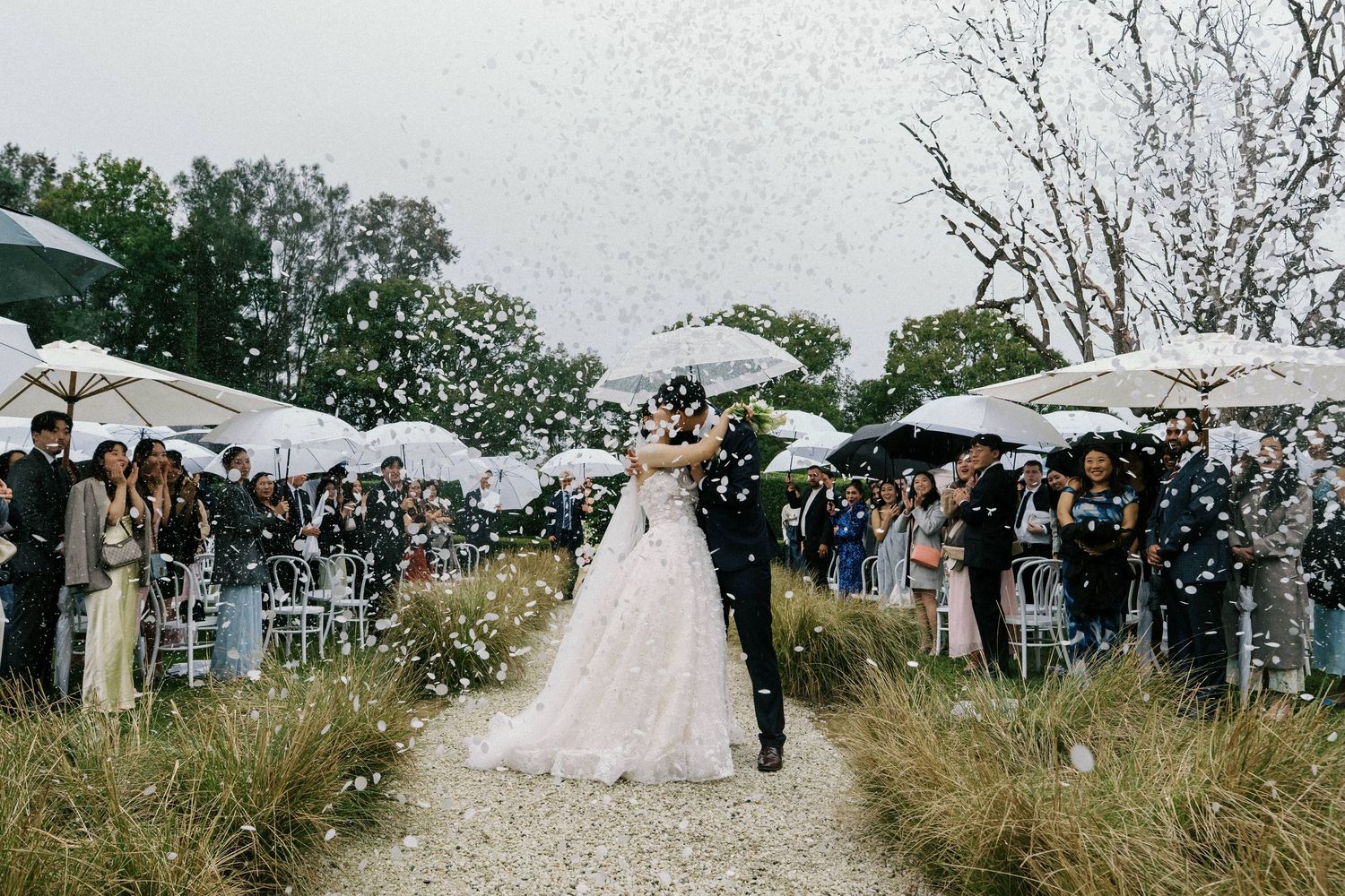 Wedding guests shower the newlyweds with confetti as they walk down a path lined with white umbrellas and tents.