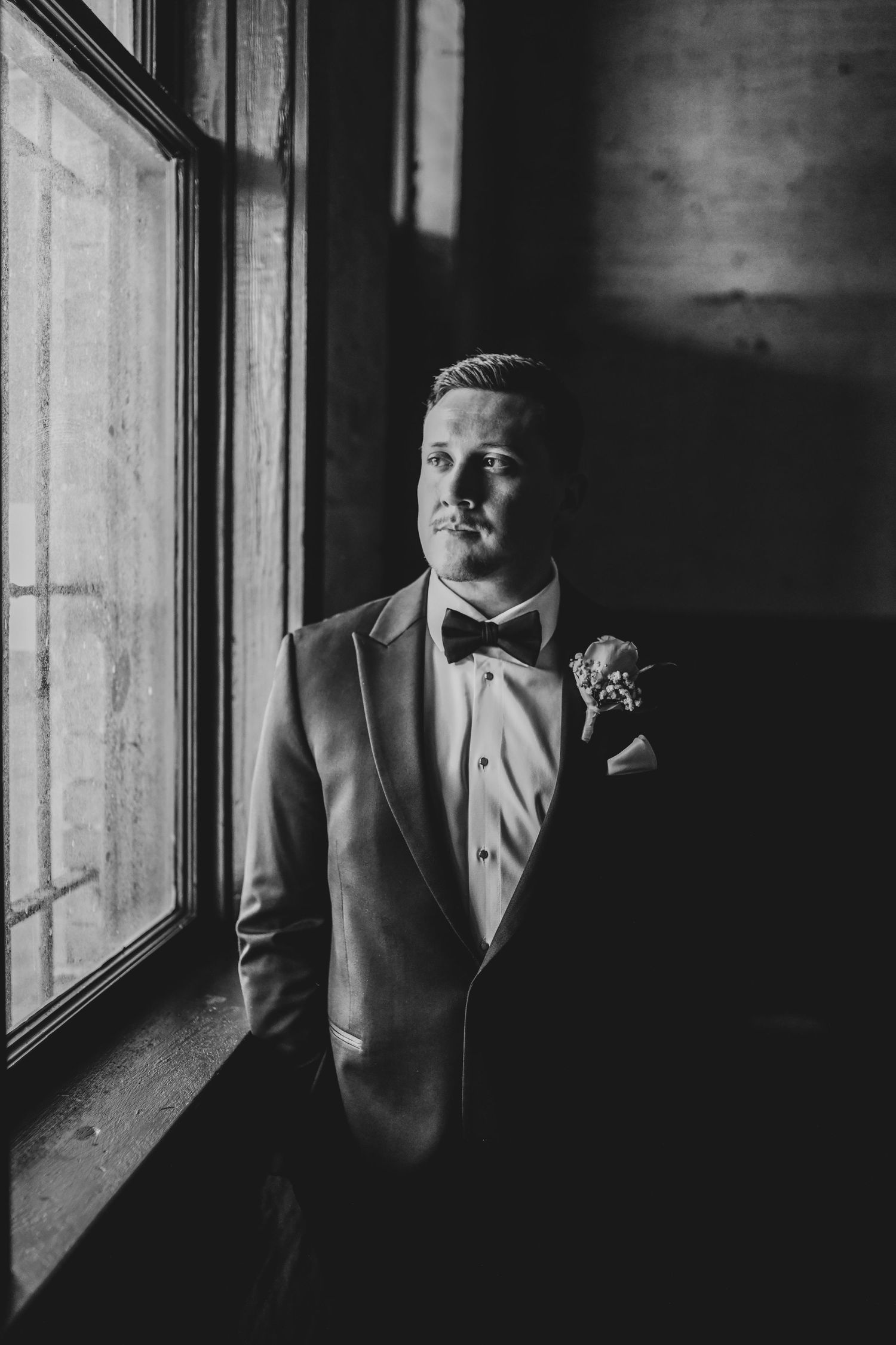 Groom standing by a large industrial window at Brake & Clutch, soft natural light highlighting his modern, editorial wedding style.