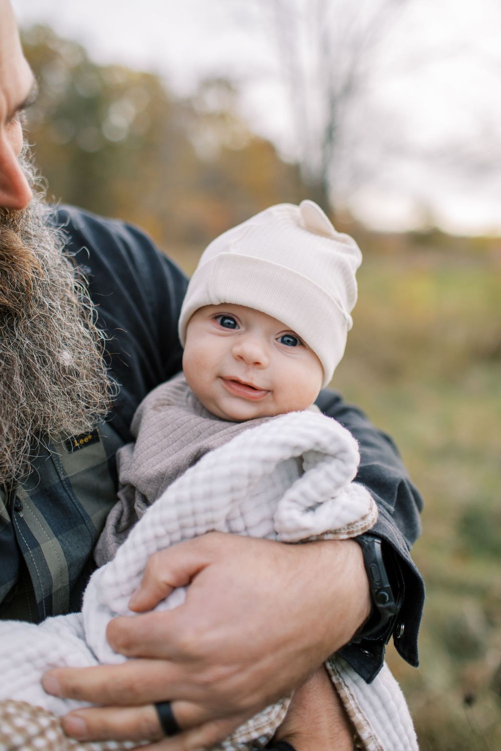 A baby wearing a white knit hat and gray sweater is held in someone's arms outdoors during autumn.