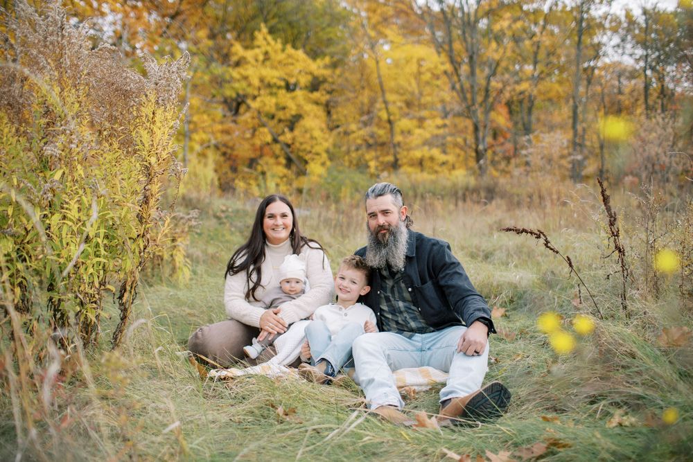 A family sits together on fallen leaves during an autumn photoshoot in a wooded area with golden foliage.