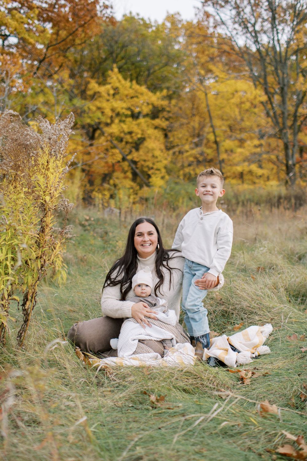 A family sits together on a blanket surrounded by golden autumn foliage and tall grass in a natural outdoor setting.