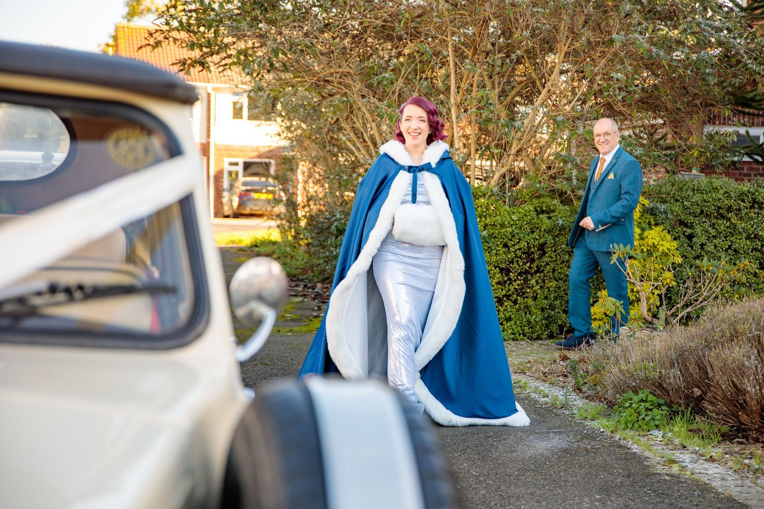 Wedding party members walk together outdoors near parked cars on an autumn day.
