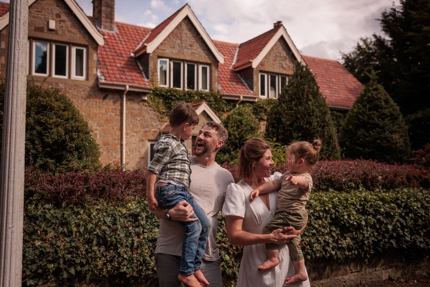 Two people playfully interact while standing in front of suburban houses with red roofs and green hedges.