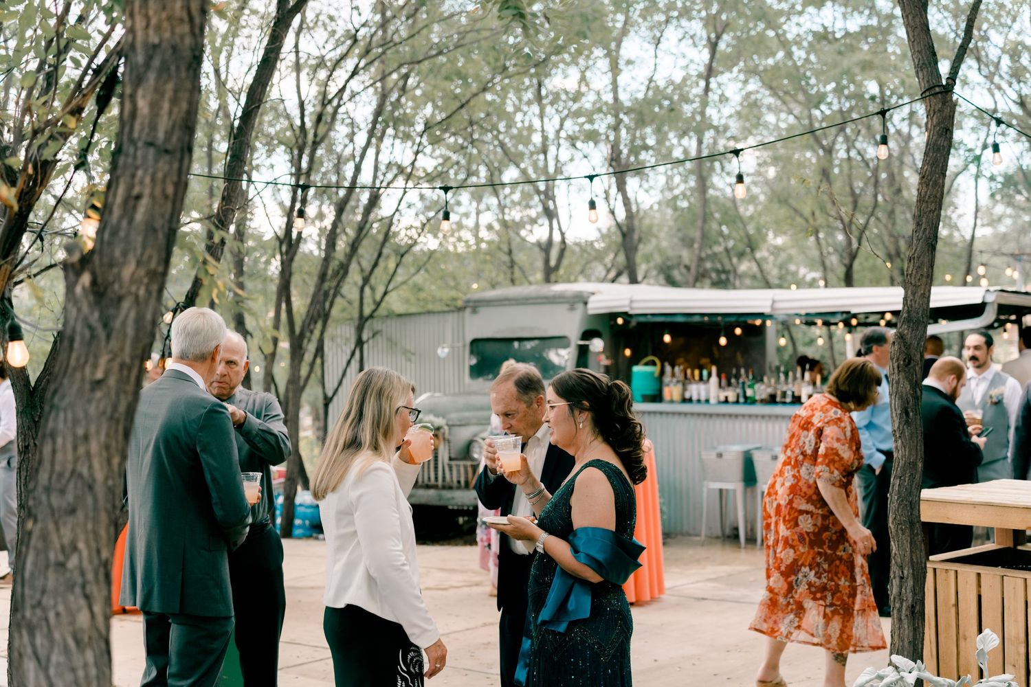 People gather at an outdoor venue with trees and string lights, socializing and enjoying drinks at a casual event.