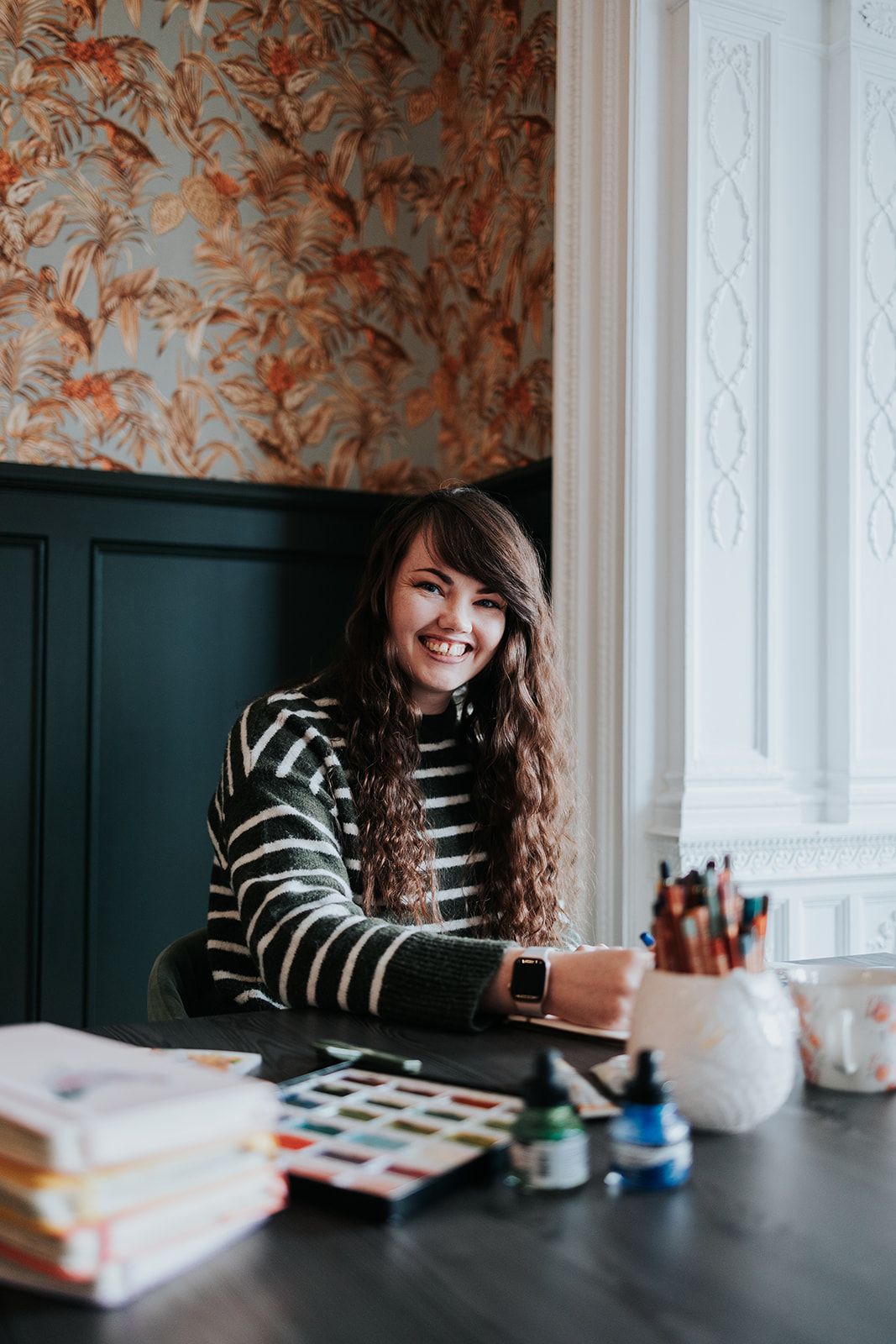 Someone in a striped sweater sits at a dark table with art supplies and books in a room with floral wallpaper.