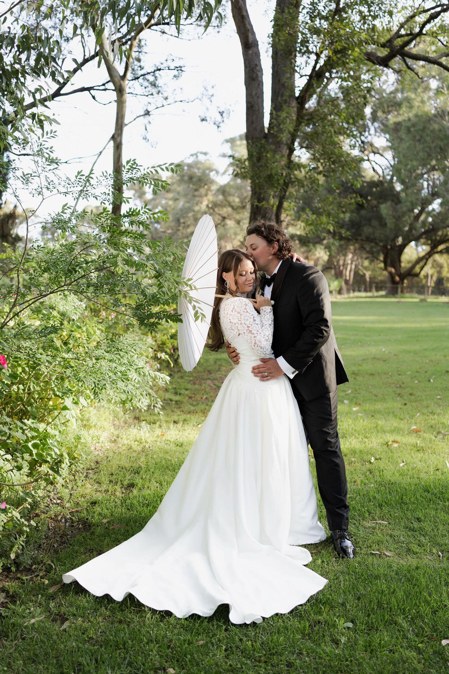 A romantic wedding couple shares a tender moment under trees with a white parasol in a lush garden setting.