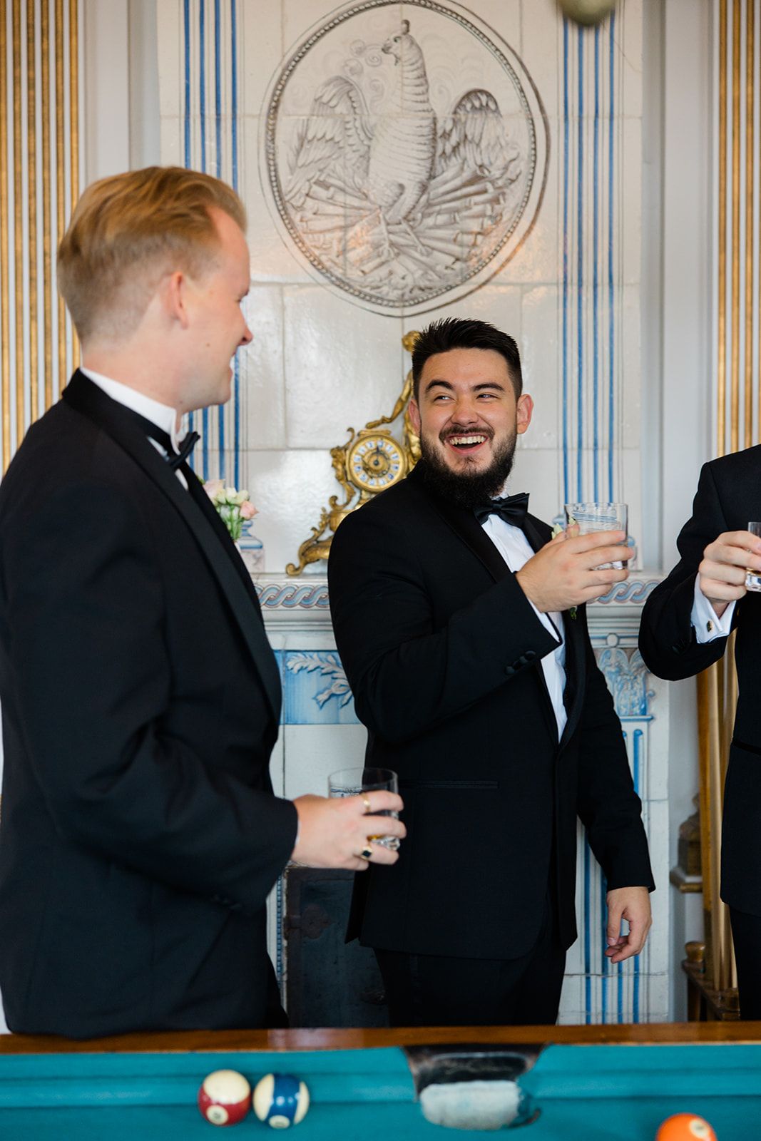 Two people in formal black tuxedos laughing and conversing while standing near a pool table.