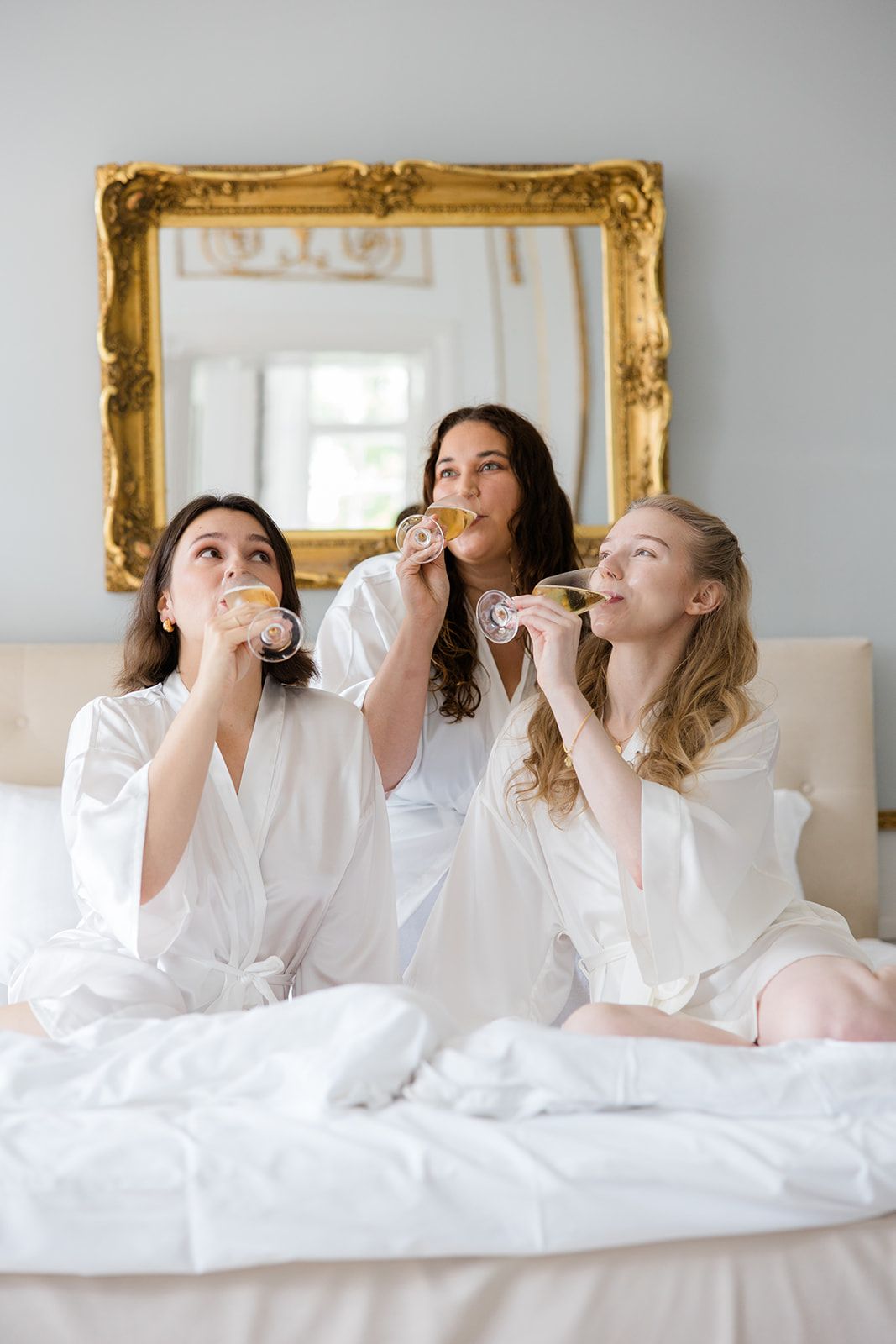 A sequence of white-robed figures lying in bed taking sips from champagne glasses in an elegant room with gold-framed mirror.