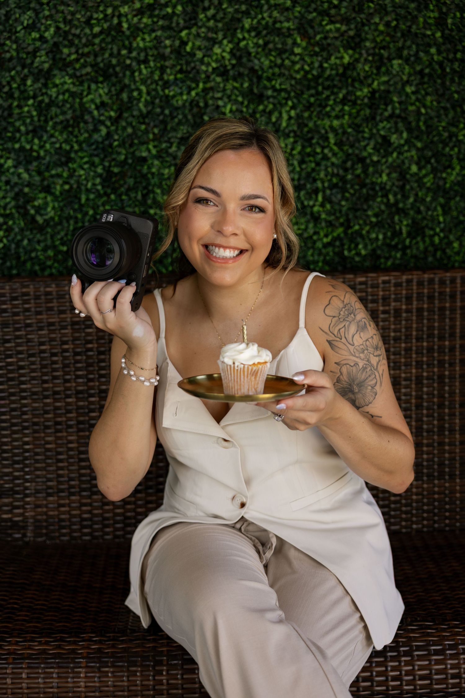 Smiling woman in a cream sleeveless outfit sits on a wicker bench holding a Cannon camera and a cupcake with a lit candle. Clearwater, FL.