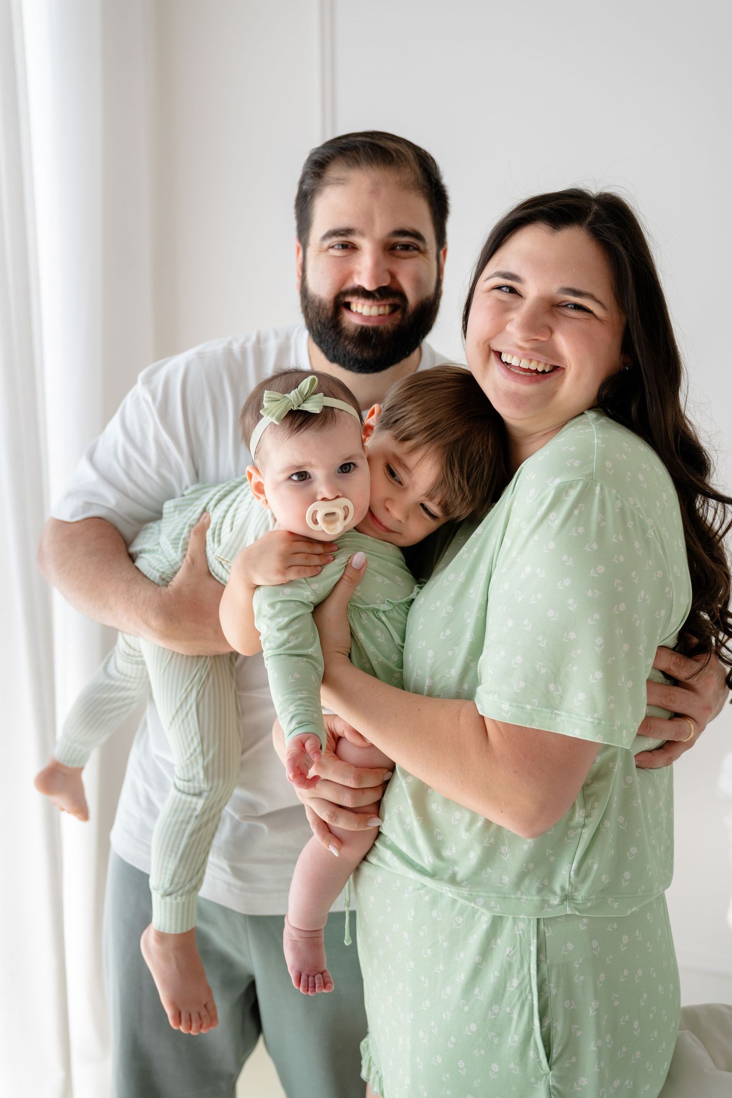 Happy family of four in green outfits, parents holding toddler and baby girl near bright window.