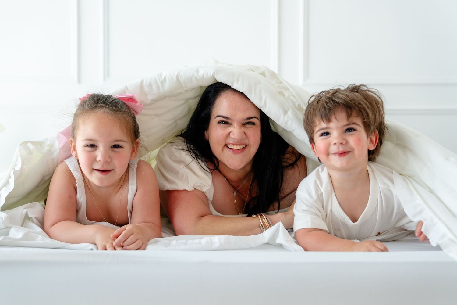 A smiling mother and two young children lying under a white duvet on a bed in a bright white room.