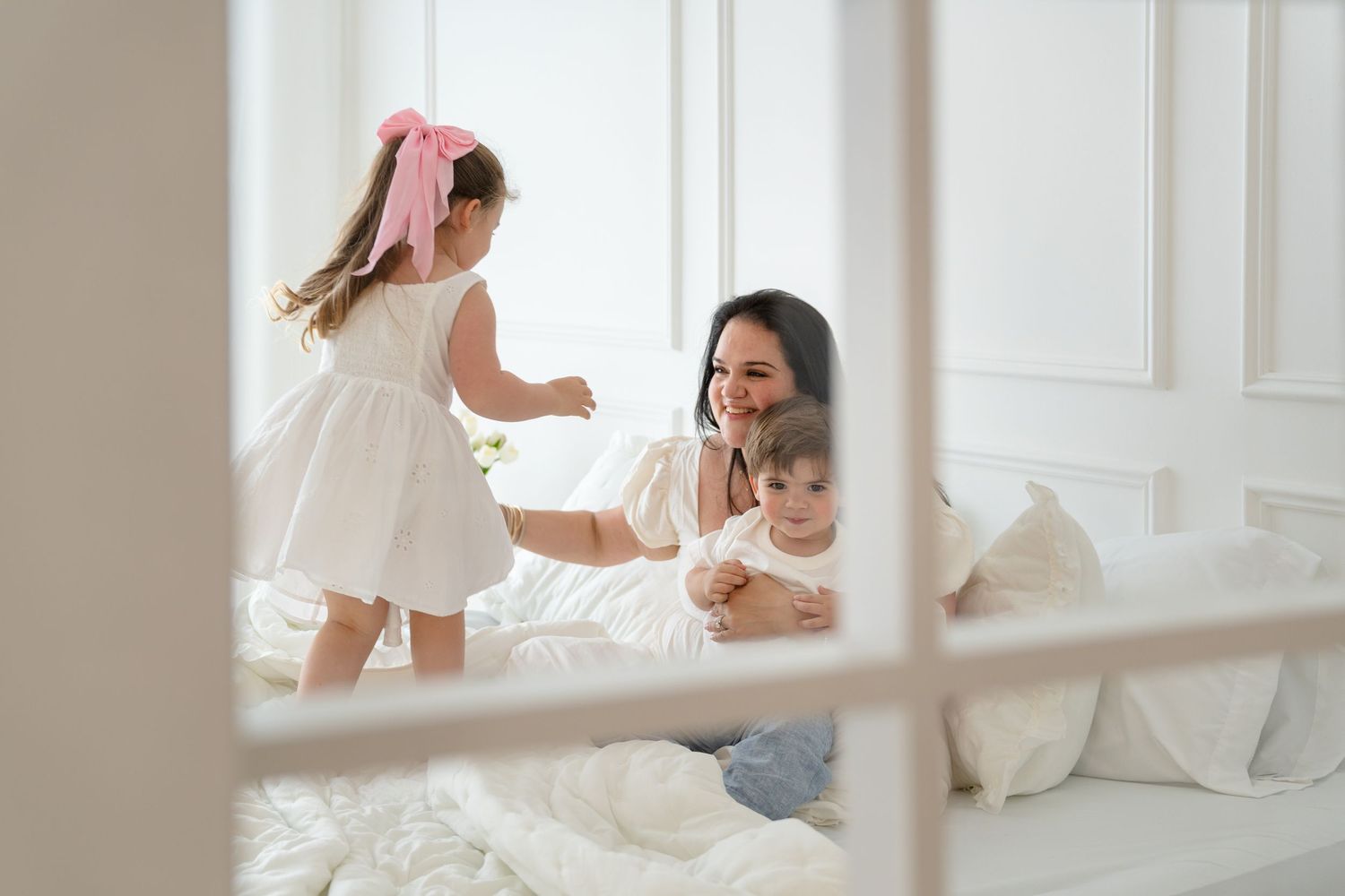 Mother smiles at baby on white bed while little girl in pink bow and white dress plays nearby.