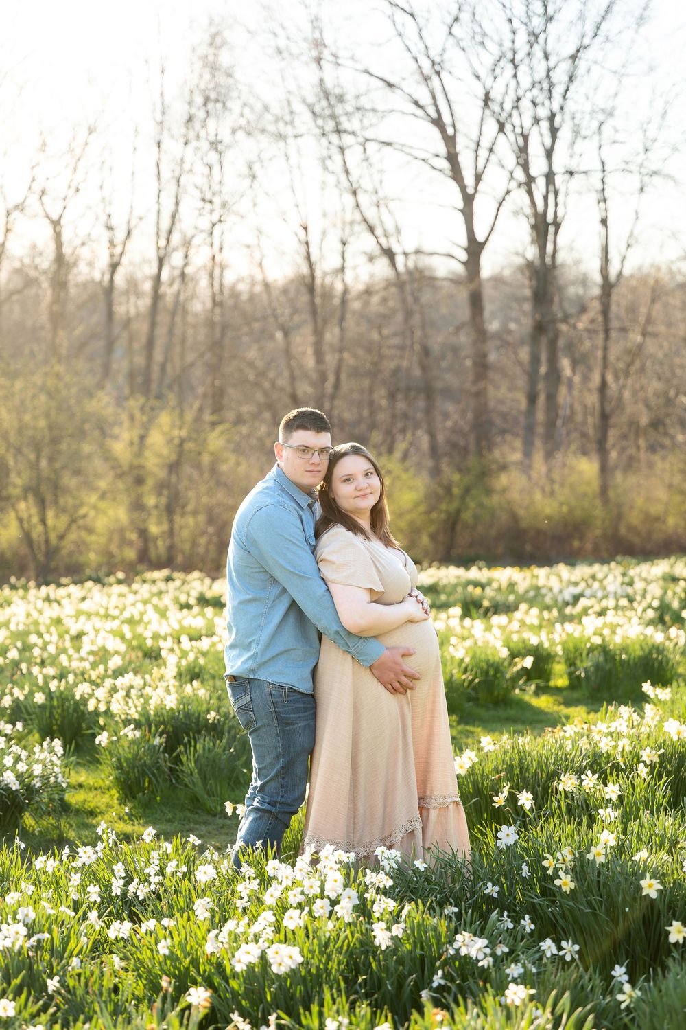 Expecting couple embracing in a sunlit field of white daffodils during spring.
