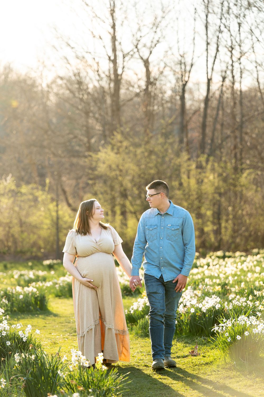 Expecting couple holding hands and walking through a sunlit spring daffodil field.