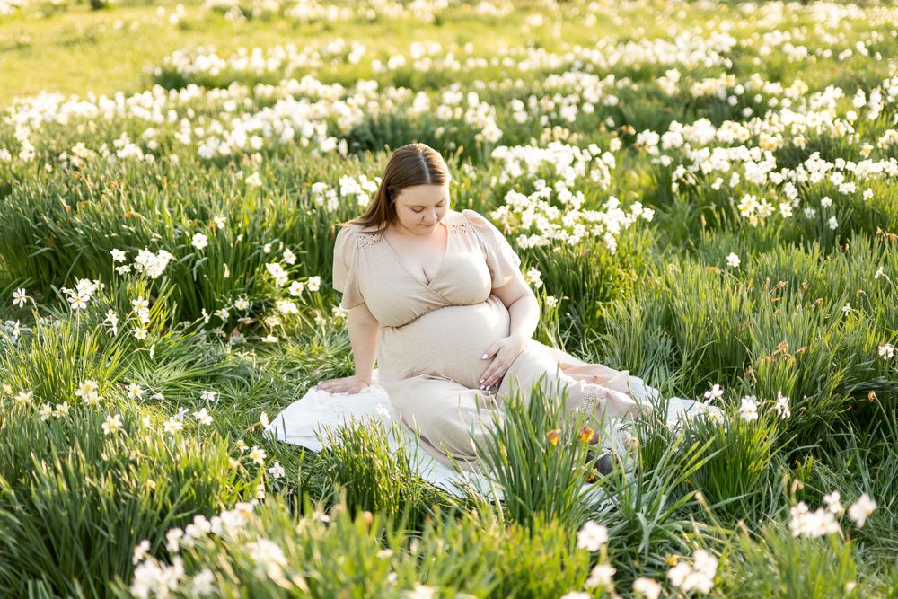 Pregnant woman in beige dress sitting among white daffodils in a sunny meadow.