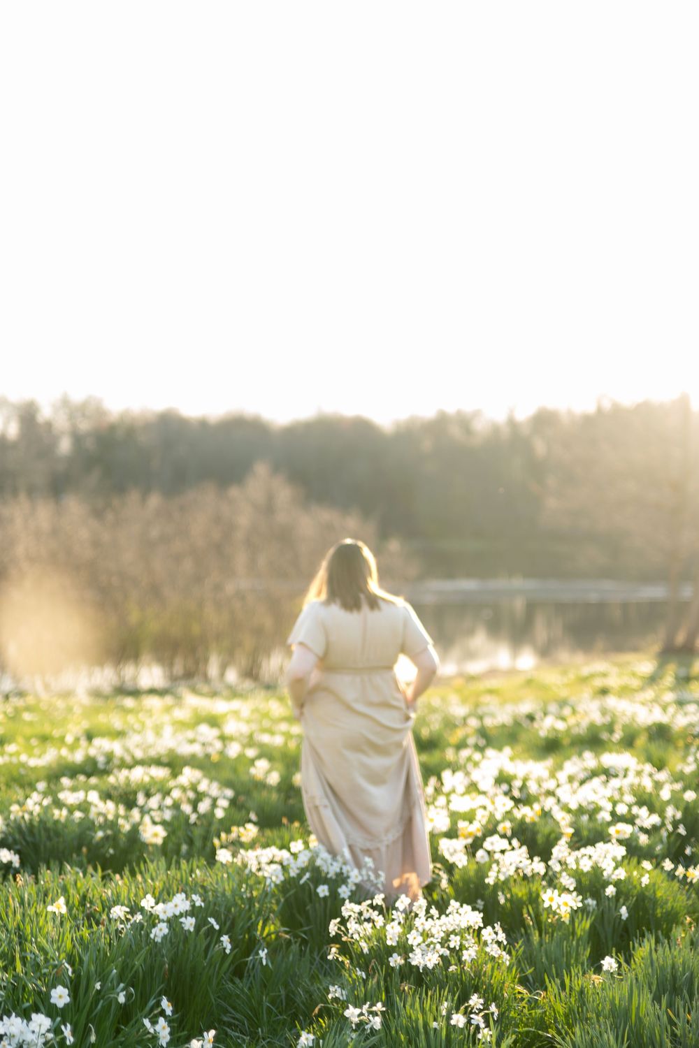 Pregnant woman in beige dress walking through a glowing field of white daffodils.