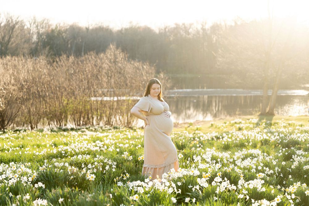 Pregnant woman in pink dress standing in daffodil field near a sunlit pond.