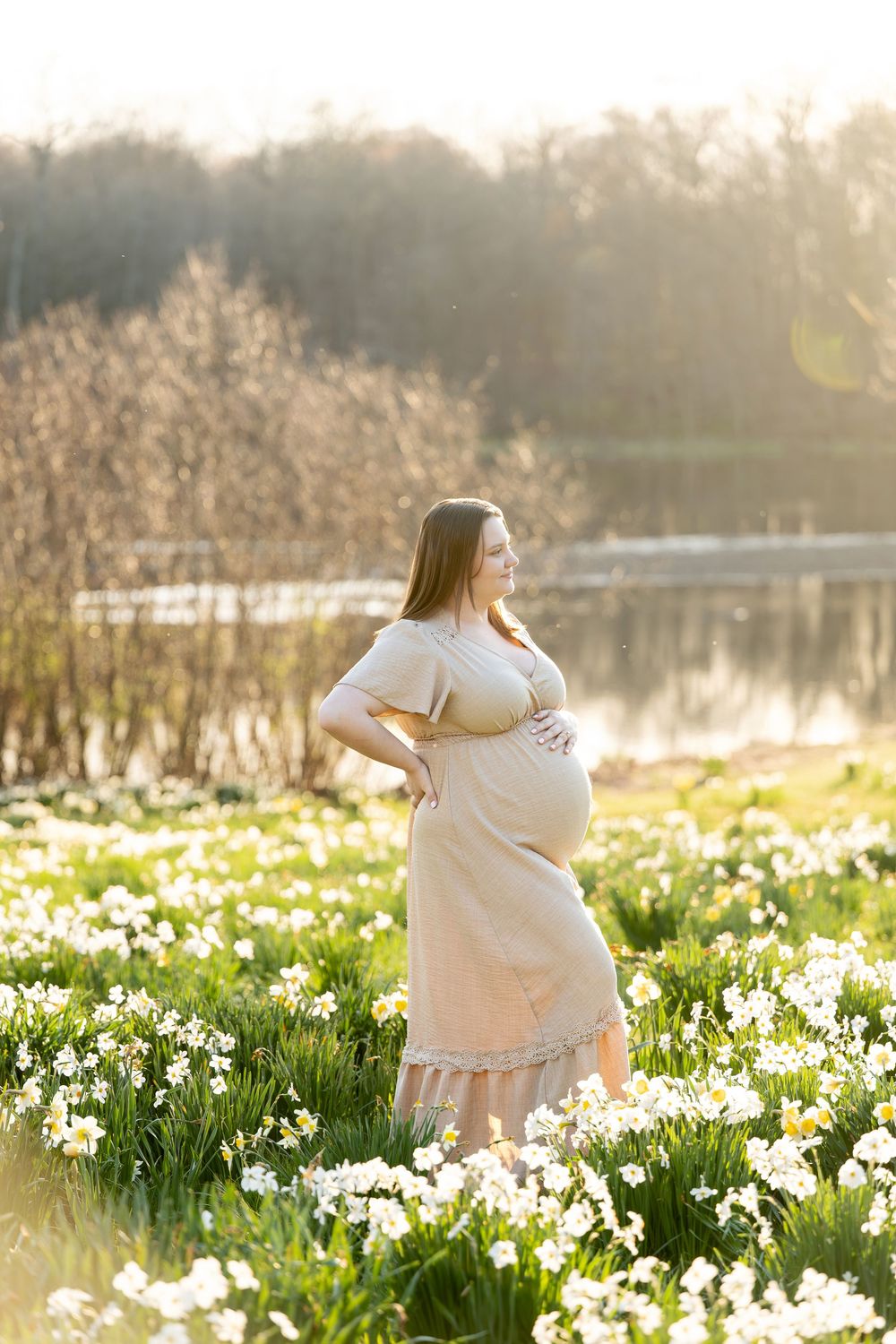 Pregnant woman in beige dress holding belly among white daffodils by a sunlit lake.