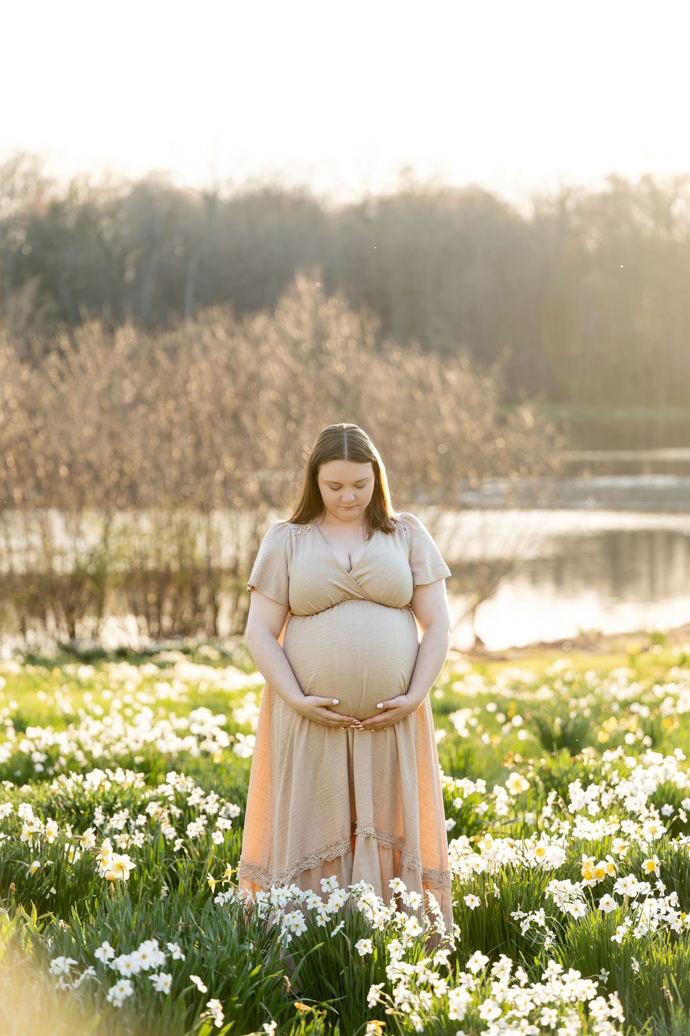 Pregnant woman in nude dress gently holding her belly surrounded by white spring flowers.