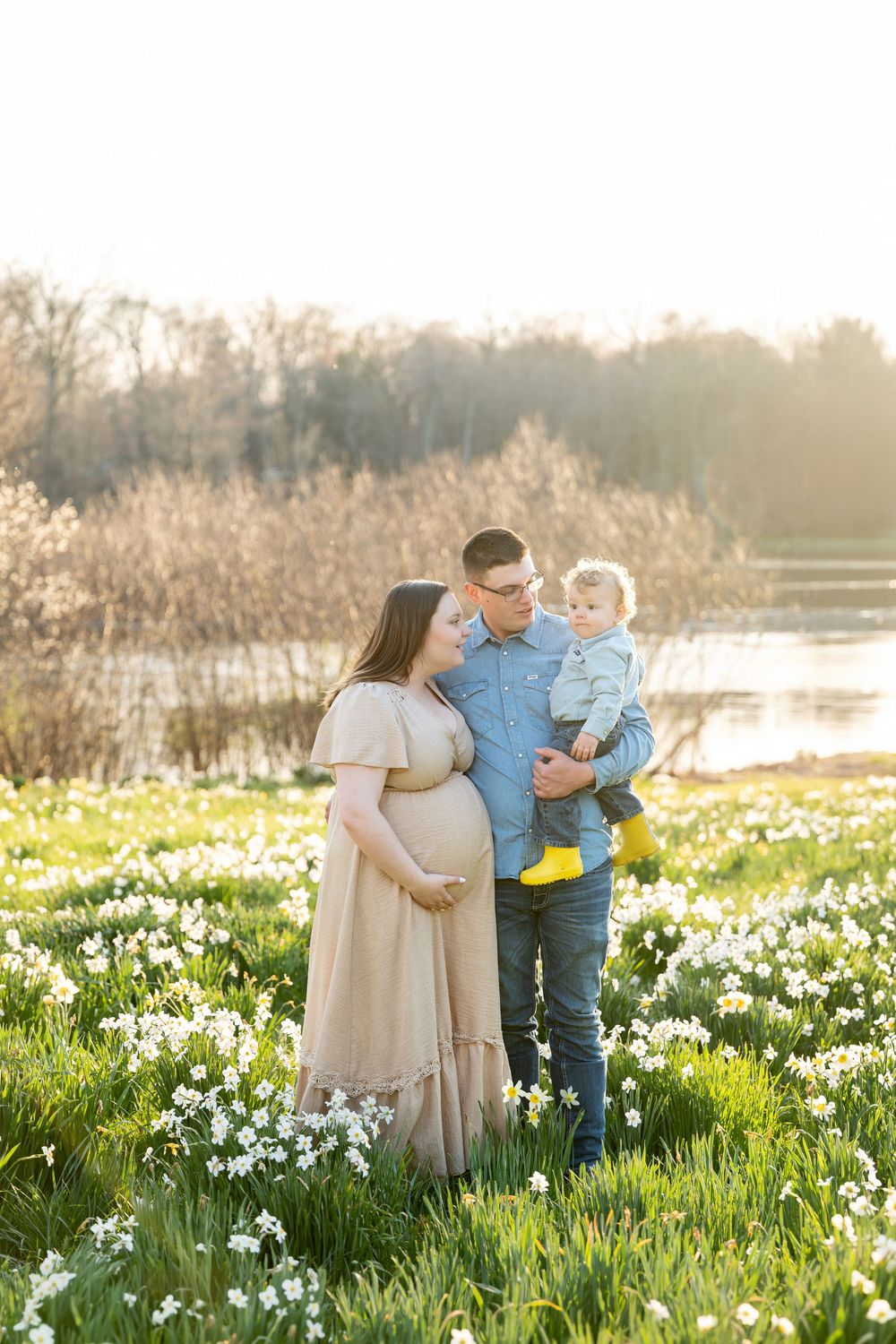 Expecting family with toddler posing in blooming daffodil field during golden hour.