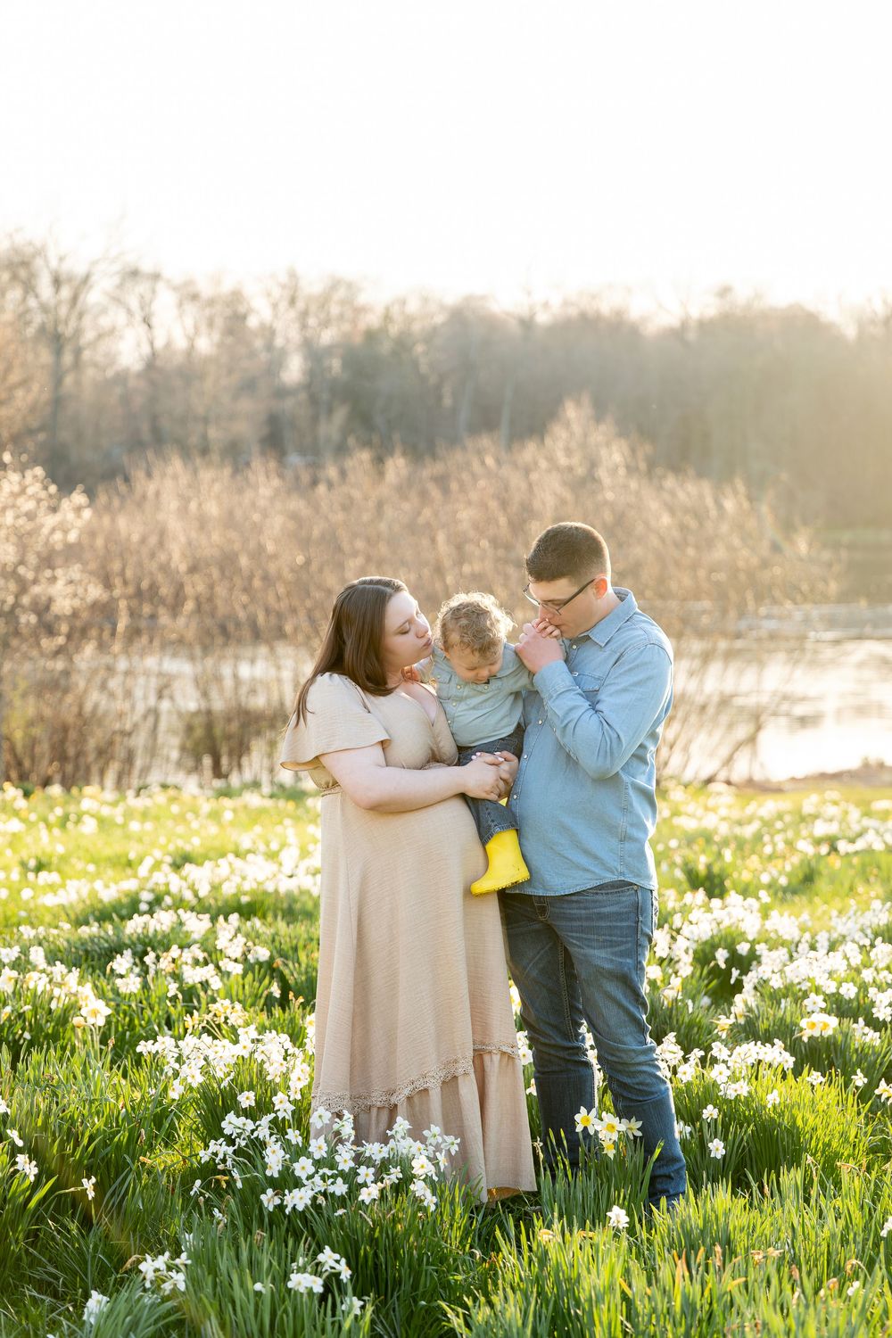 Parents kissing young child in a sunlit field of white daffodils near a lake.