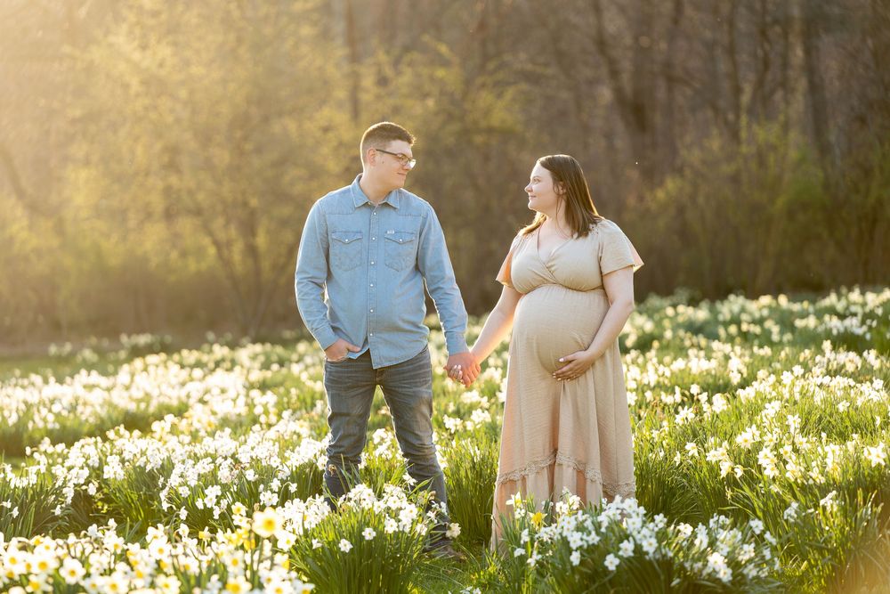 Expecting couple holding hands and smiling in a golden sunlit field of daffodils.