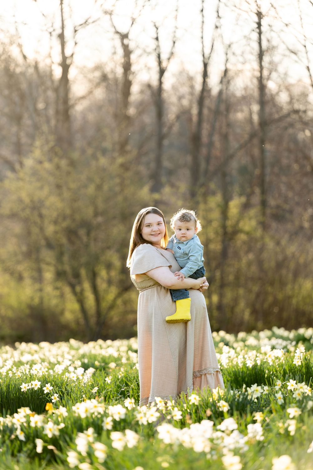 Pregnant mother in blush dress holding baby boy in yellow boots in daffodil field.