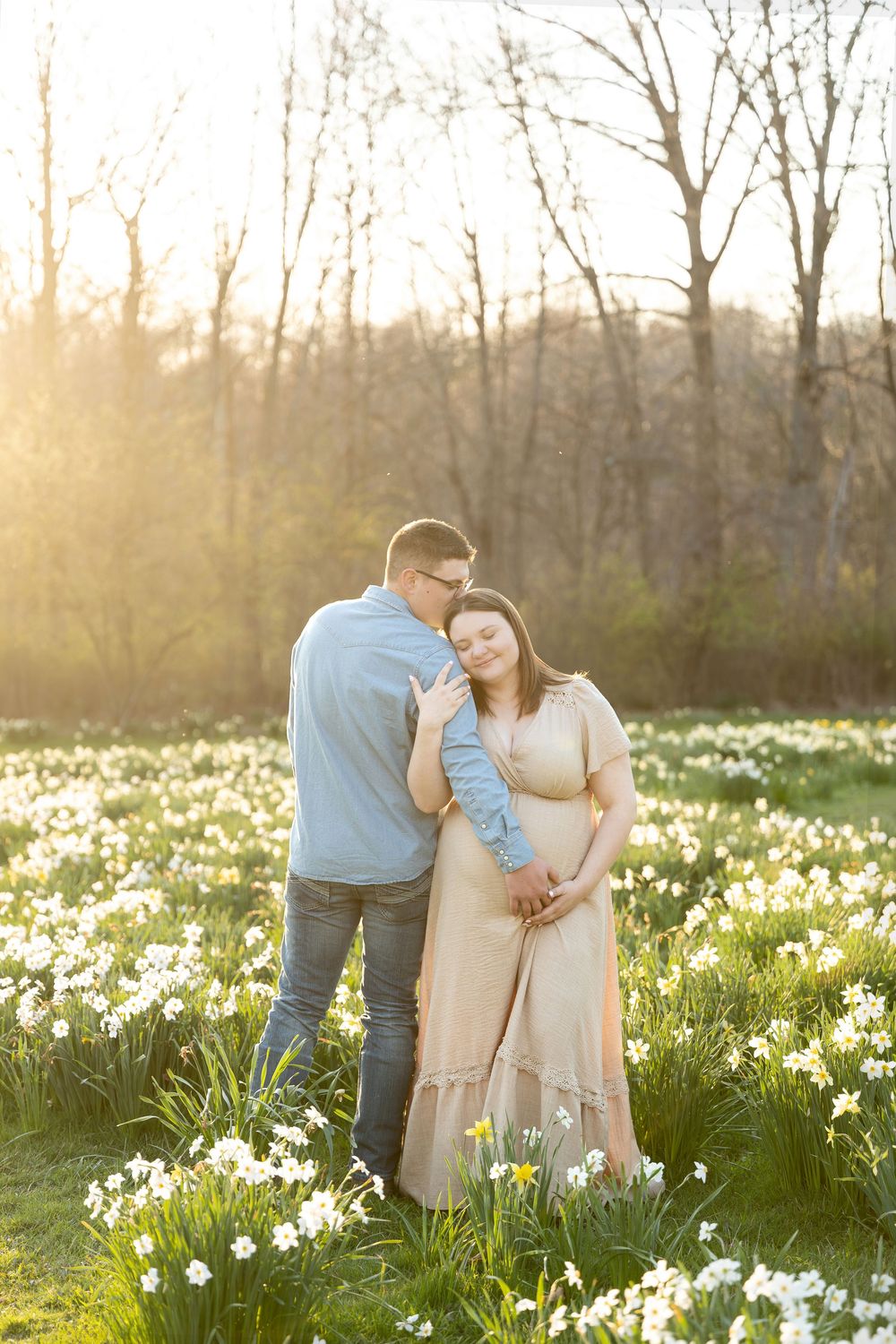Expectant couple embracing in golden hour light surrounded by blooming white daffodil field.