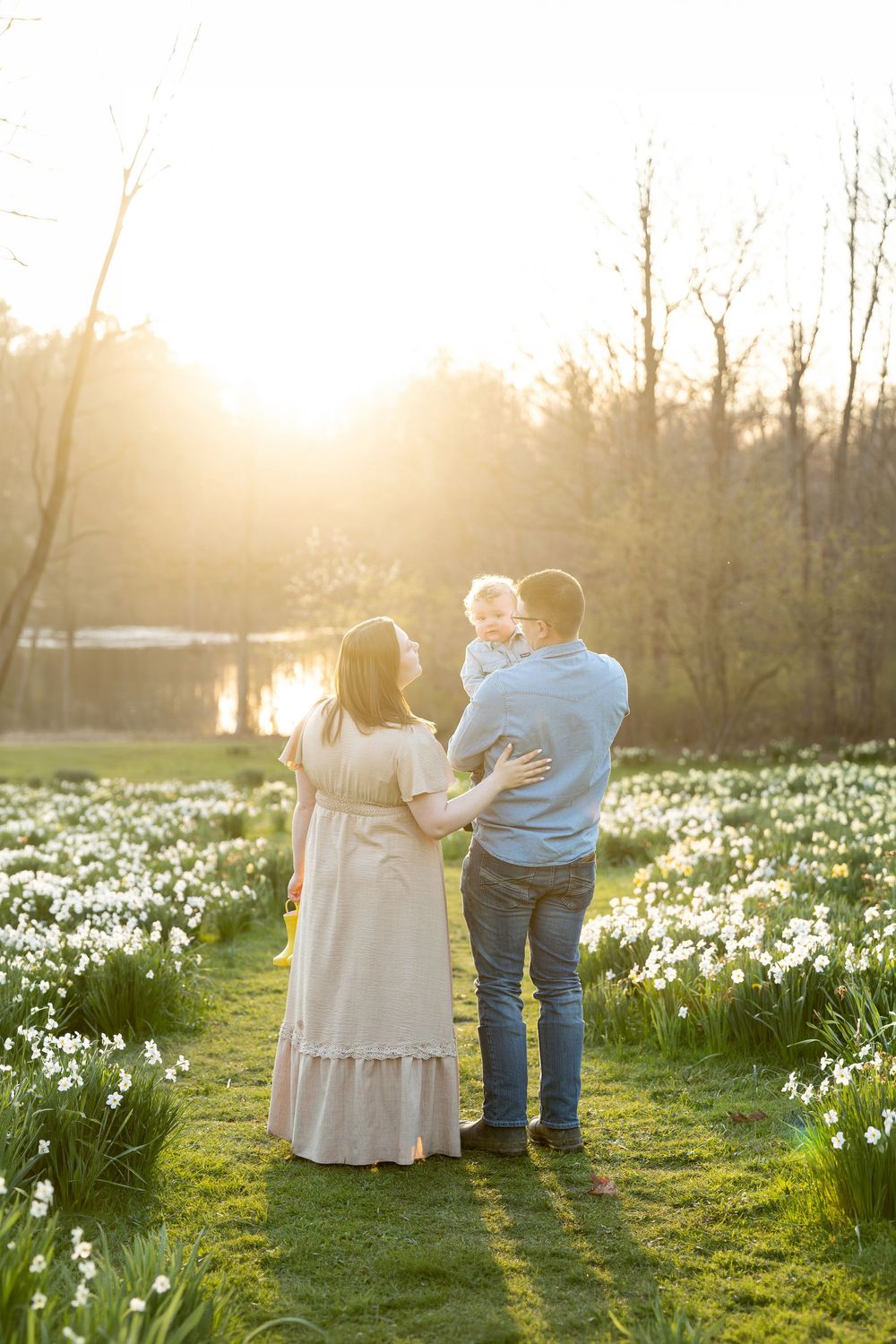 Family of three standing in daffodil field at golden sunset, father holding toddler near lake.