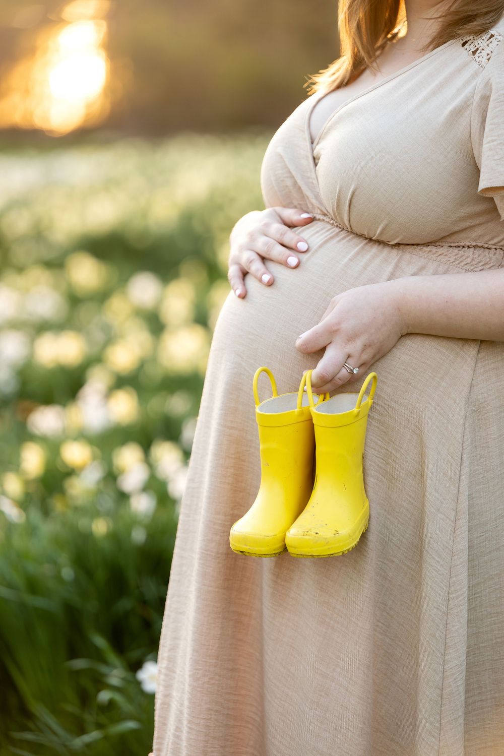 Pregnant woman in beige dress holding tiny yellow rain boots beside her baby bump in flower field.