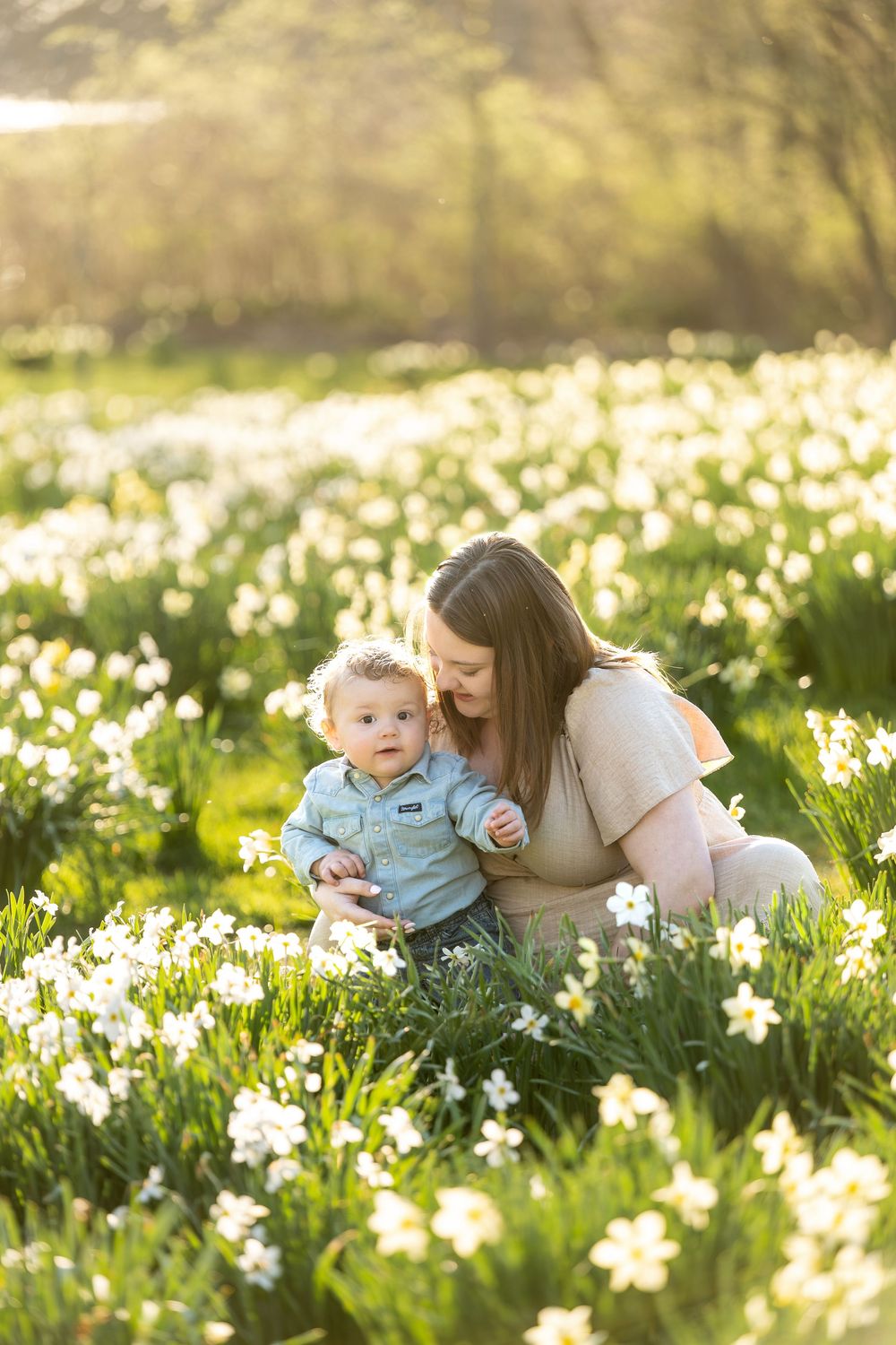 Pregnant mother sitting with baby boy among white daffodils in warm golden spring light.