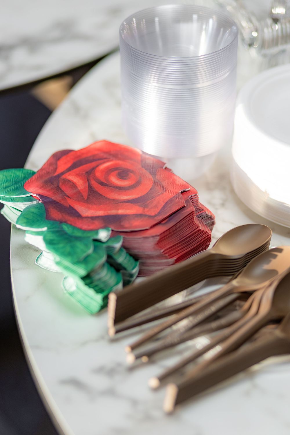 Rose-shaped napkins beside gold cutlery, clear plastic cups, and white plates on a marble table.